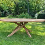 Tuintafel gemaakt van teakhout. Het blad heeft een ovale vorm. Onder het blad heeft de tafel een kruispoot